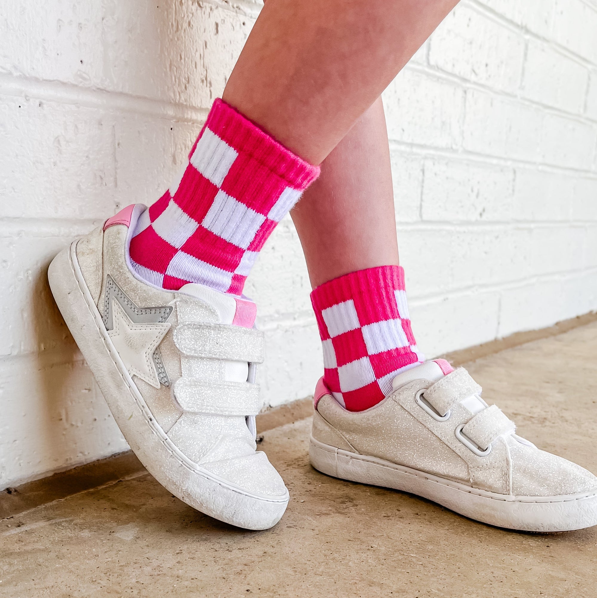 White sneakers with pink and white checkered socks against a white brick wall.