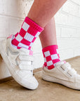 White sneakers with pink and white checkered socks against a white brick wall.