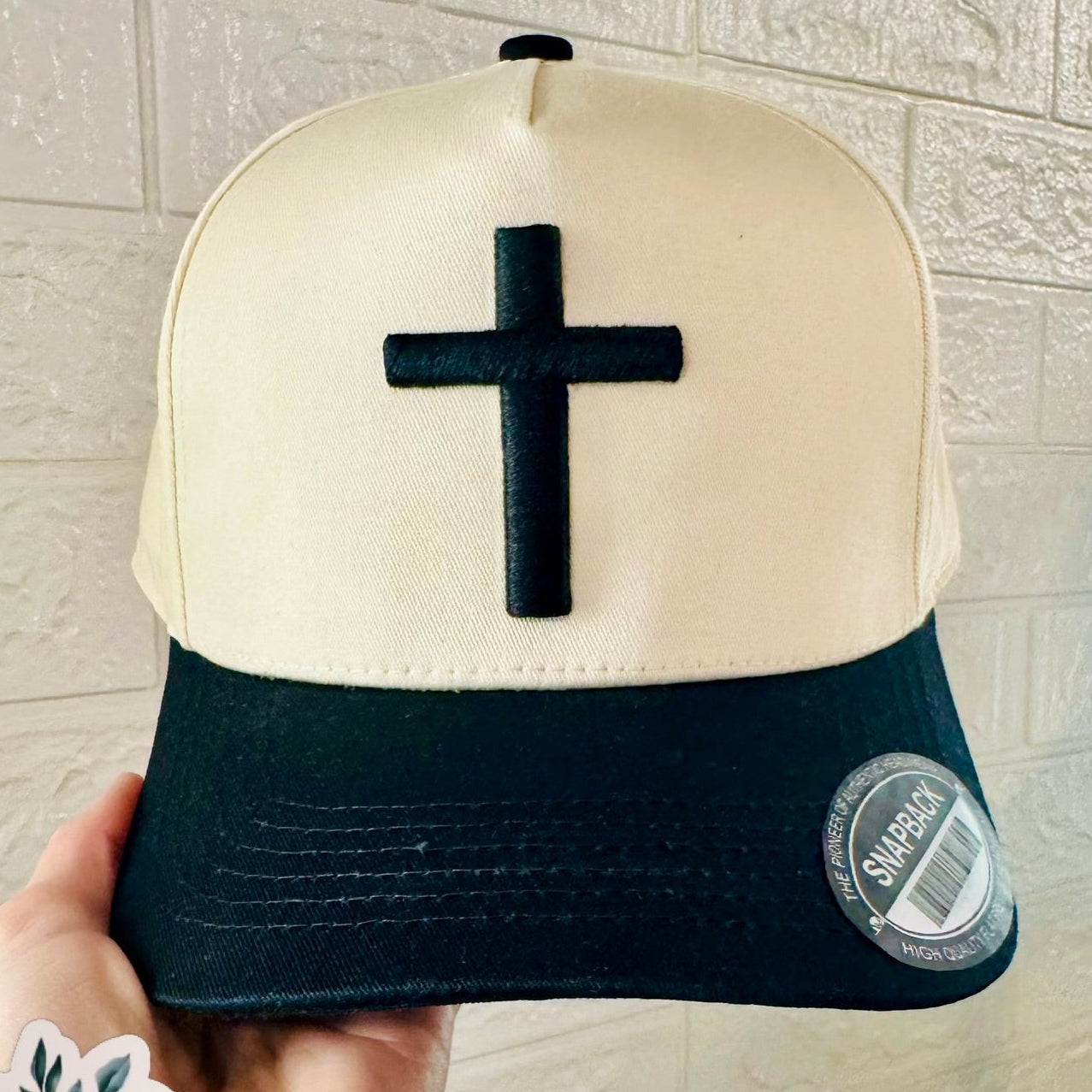 Beige and navy cap with black cross held against a light brick wall.