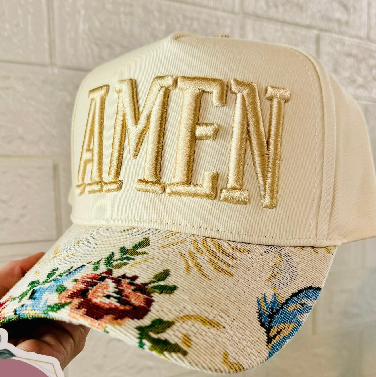 Beige cap with floral embroidery and 'AMEN' text, held by a hand against a light background.