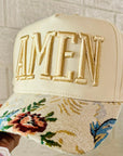 Beige cap with floral embroidery and 'AMEN' text, held by a hand against a light background.