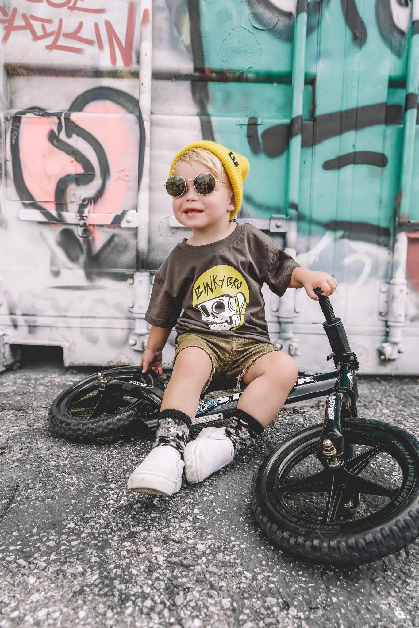 Child sitting on a balance bike in front of a graffiti-covered wall