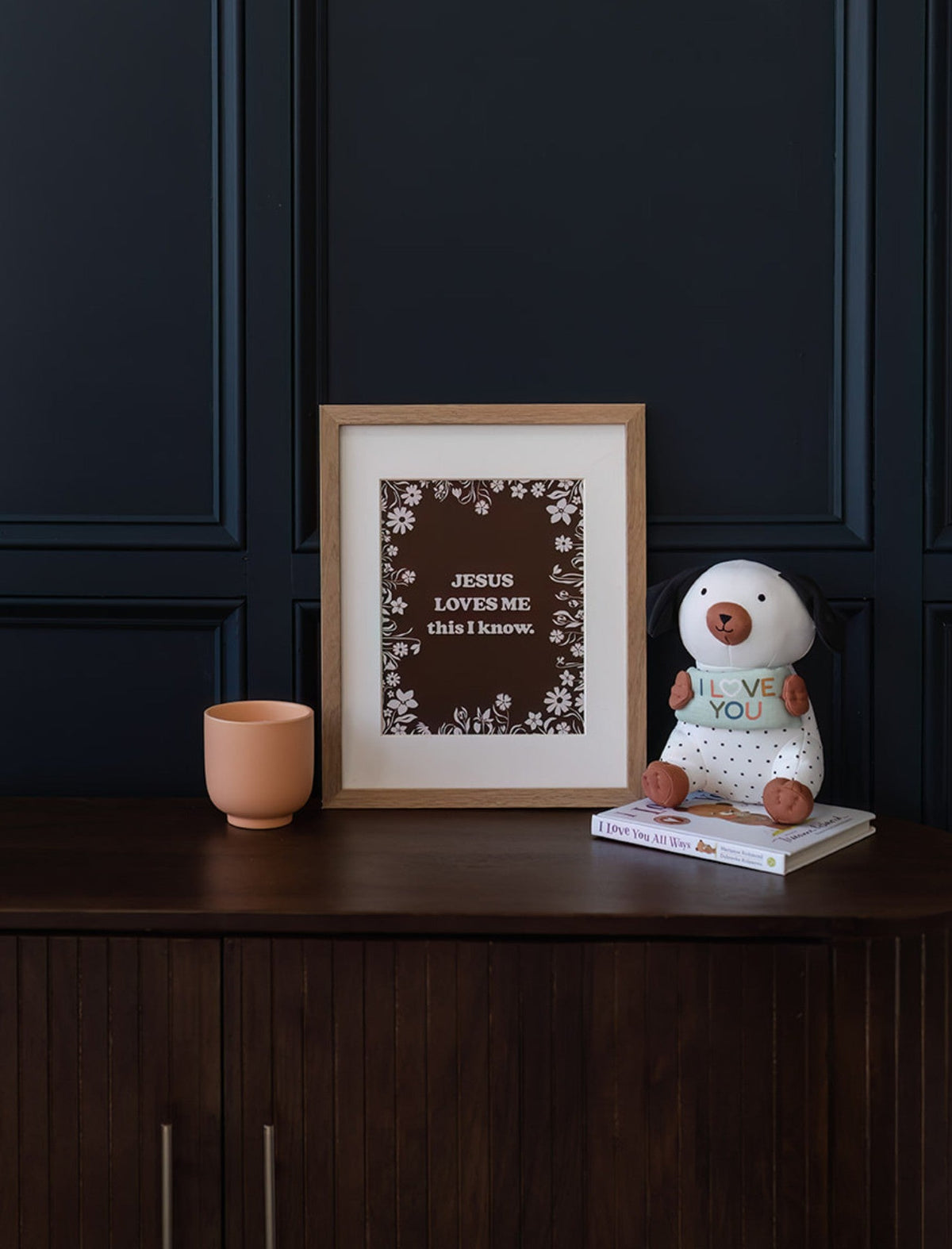 Framed Jesus Loves Me floral art print displayed on a dark wooden cabinet beside a cup and decorative figure