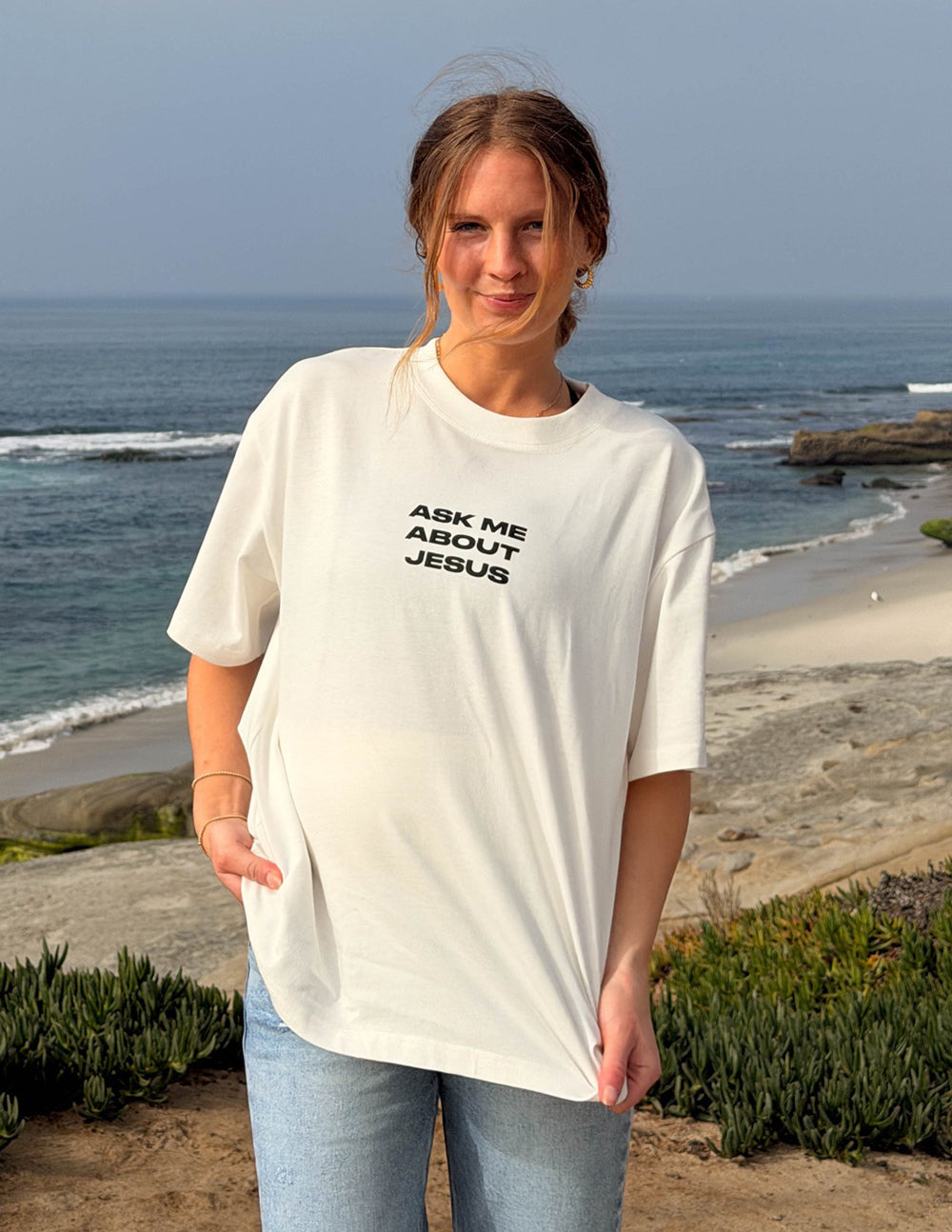 Model wearing cream Ask Me About Jesus graphic tee by the coast
