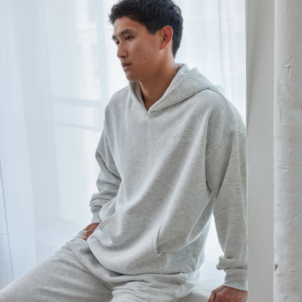 Person wearing a gray hoodie and pants sitting against a white curtain.