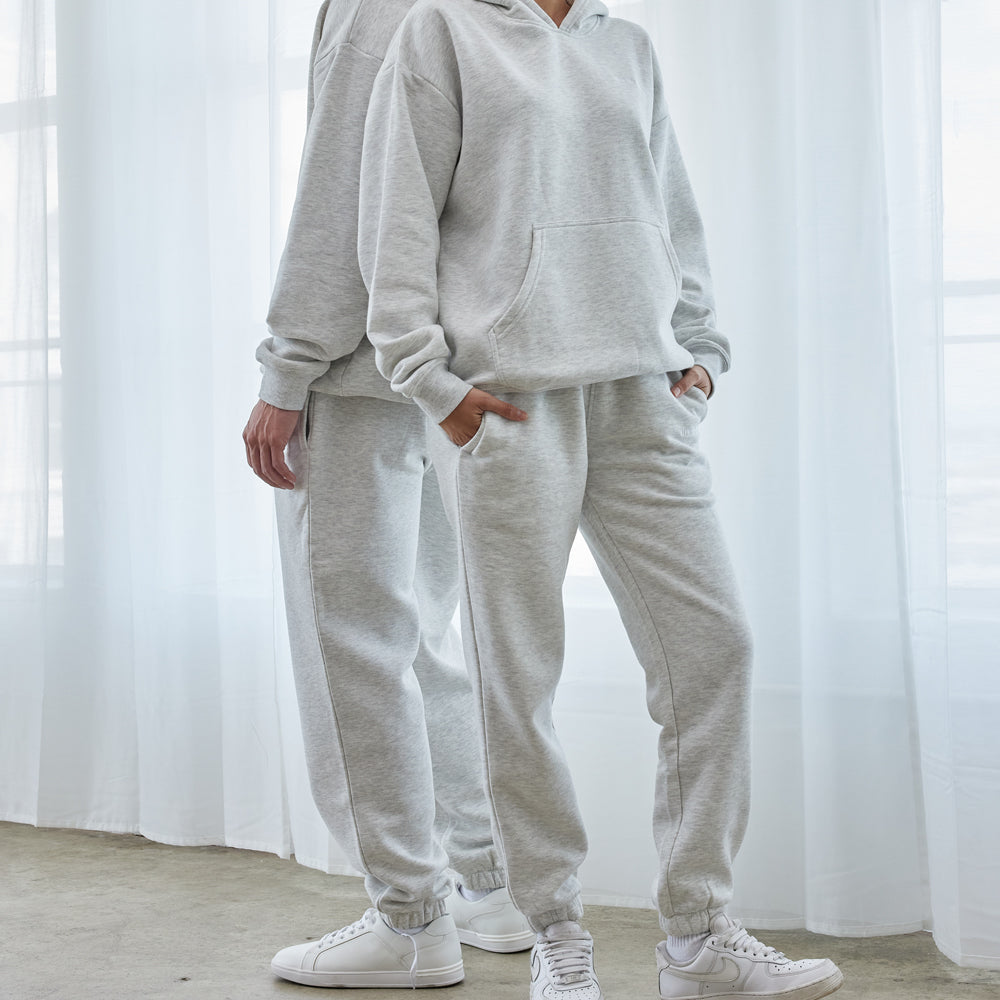 Two people wearing matching gray tracksuits standing in a bright room with white curtains.