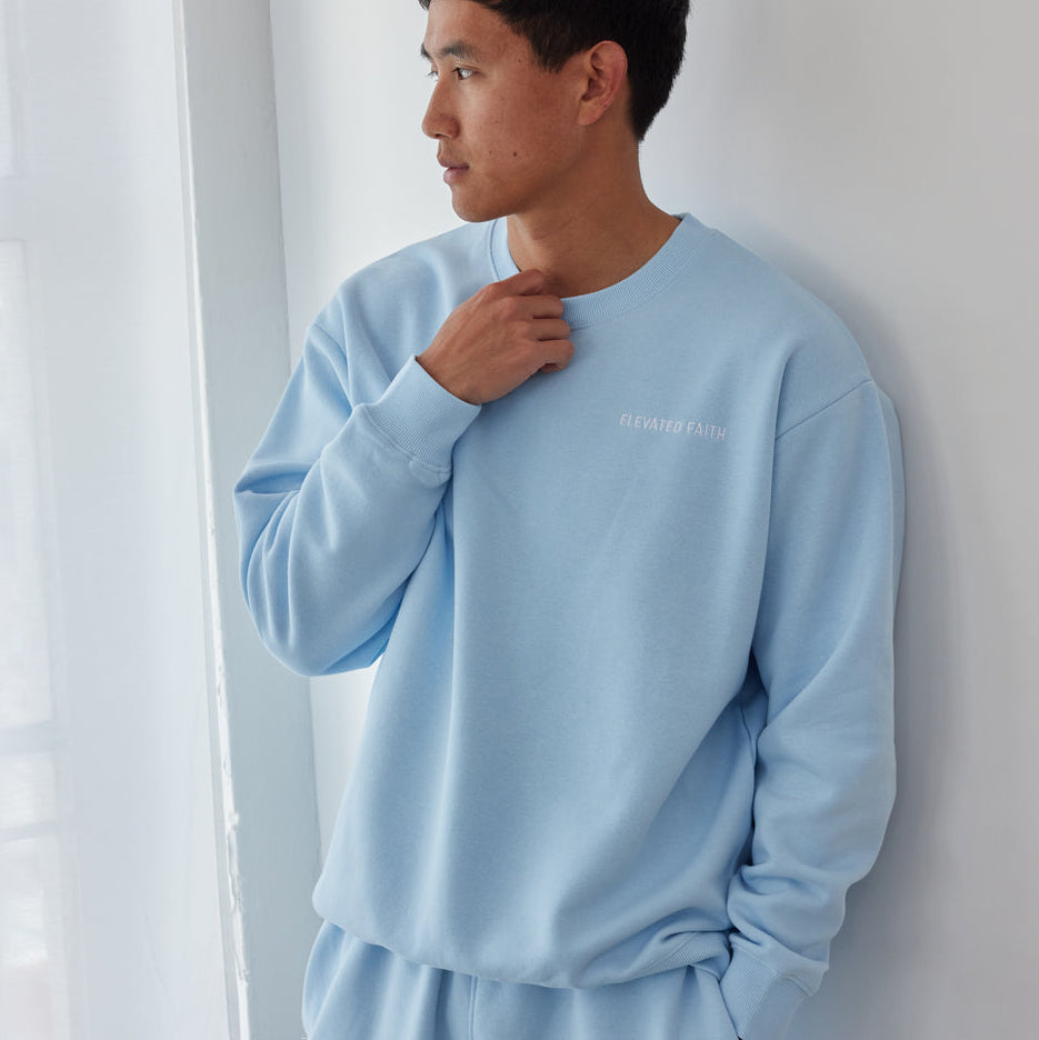 Person wearing a light blue sweatshirt and pants set against a white curtain.