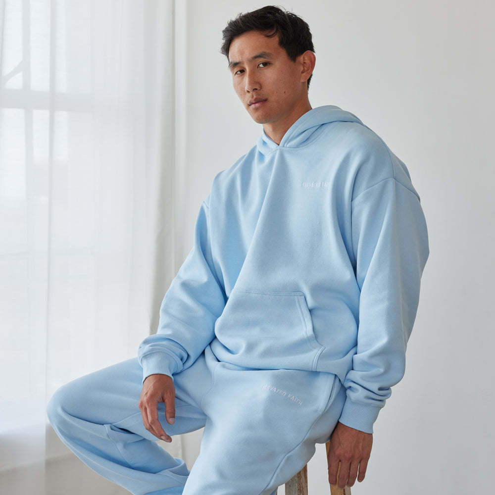 Person wearing a light blue tracksuit sitting on a wooden stool in a minimalistic room.