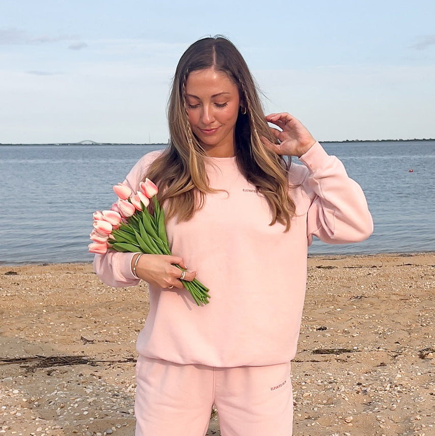Woman in pink outfit holding greenery on a beach with ocean in the background