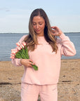 Woman in pink outfit holding greenery on a beach with ocean in the background