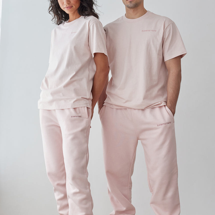 Two people wearing matching light pink outfits standing against a white wall.