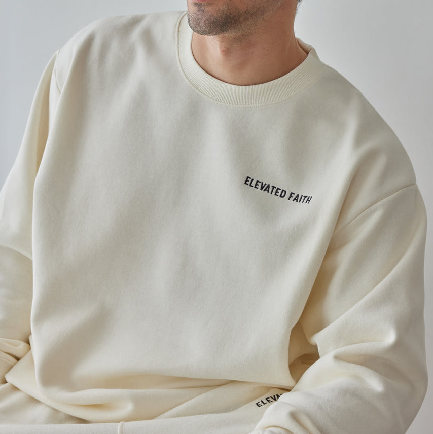 Man wearing a cream-colored sweatshirt with 'Elevated Faith' text on a plain background