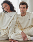 Two people wearing matching white sweatshirts and pants sitting on a light-colored floor.