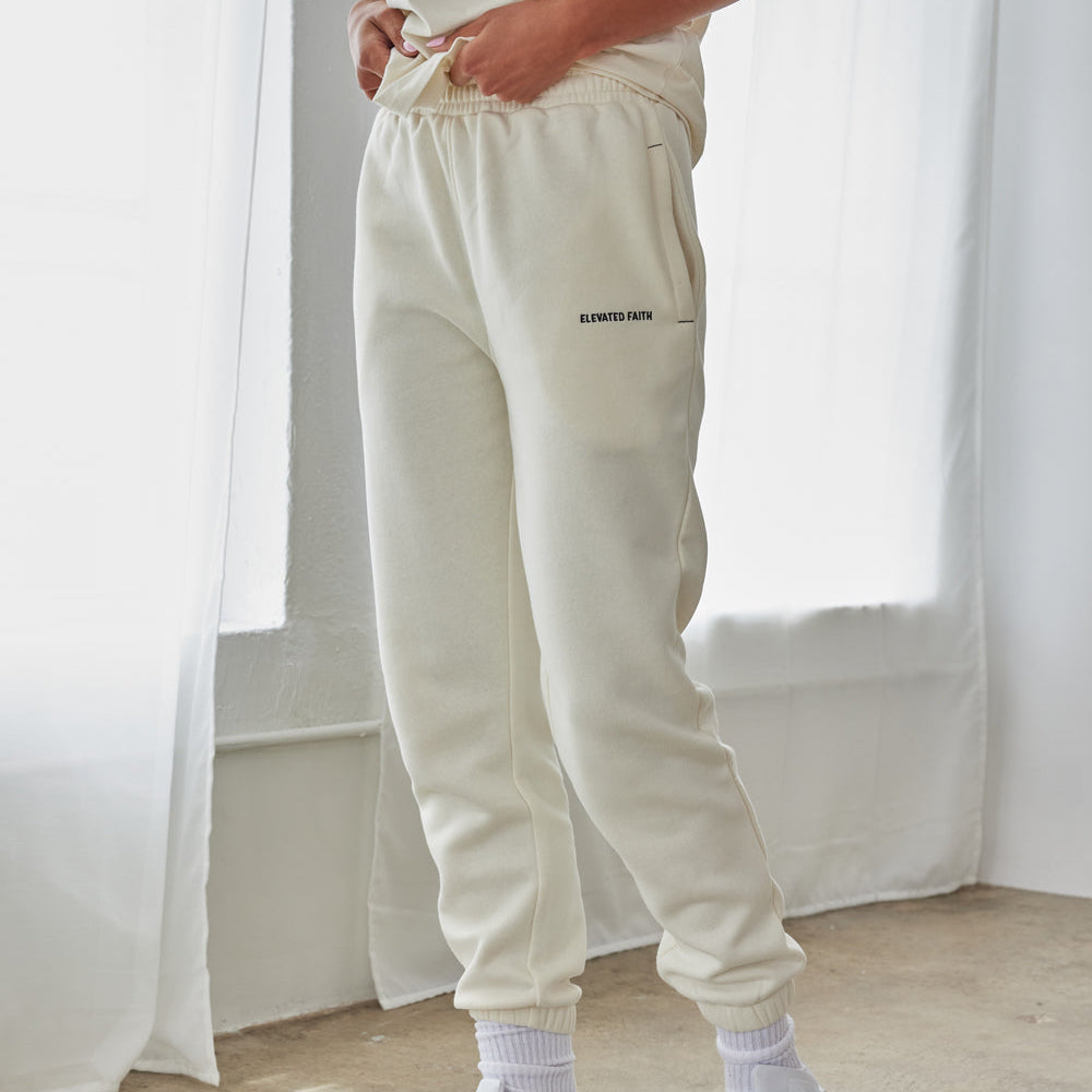 Person wearing light gray sweatpants with a brand logo, white sneakers, and socks in a minimalistic setting.
