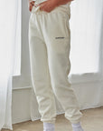 Person wearing light gray sweatpants with a brand logo, white sneakers, and socks in a minimalistic setting.