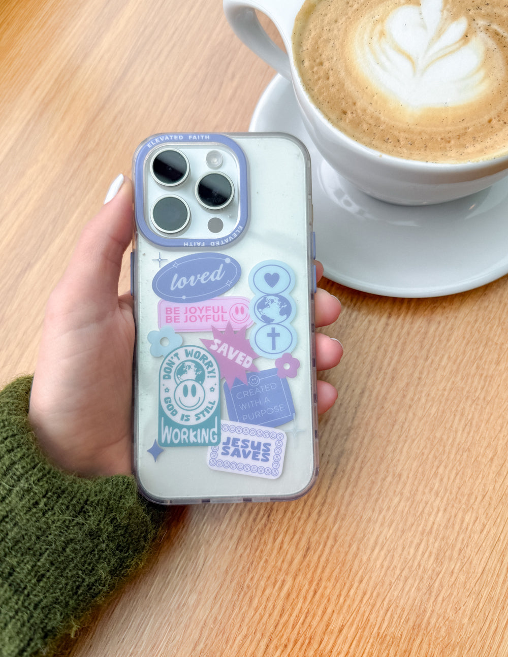 Hand holding an iPhone in the pastel Be Joyful phone case beside a cup of coffee