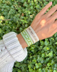 Beige WWJD bracelet styled alongside a green woven bracelet on a wrist outdoors