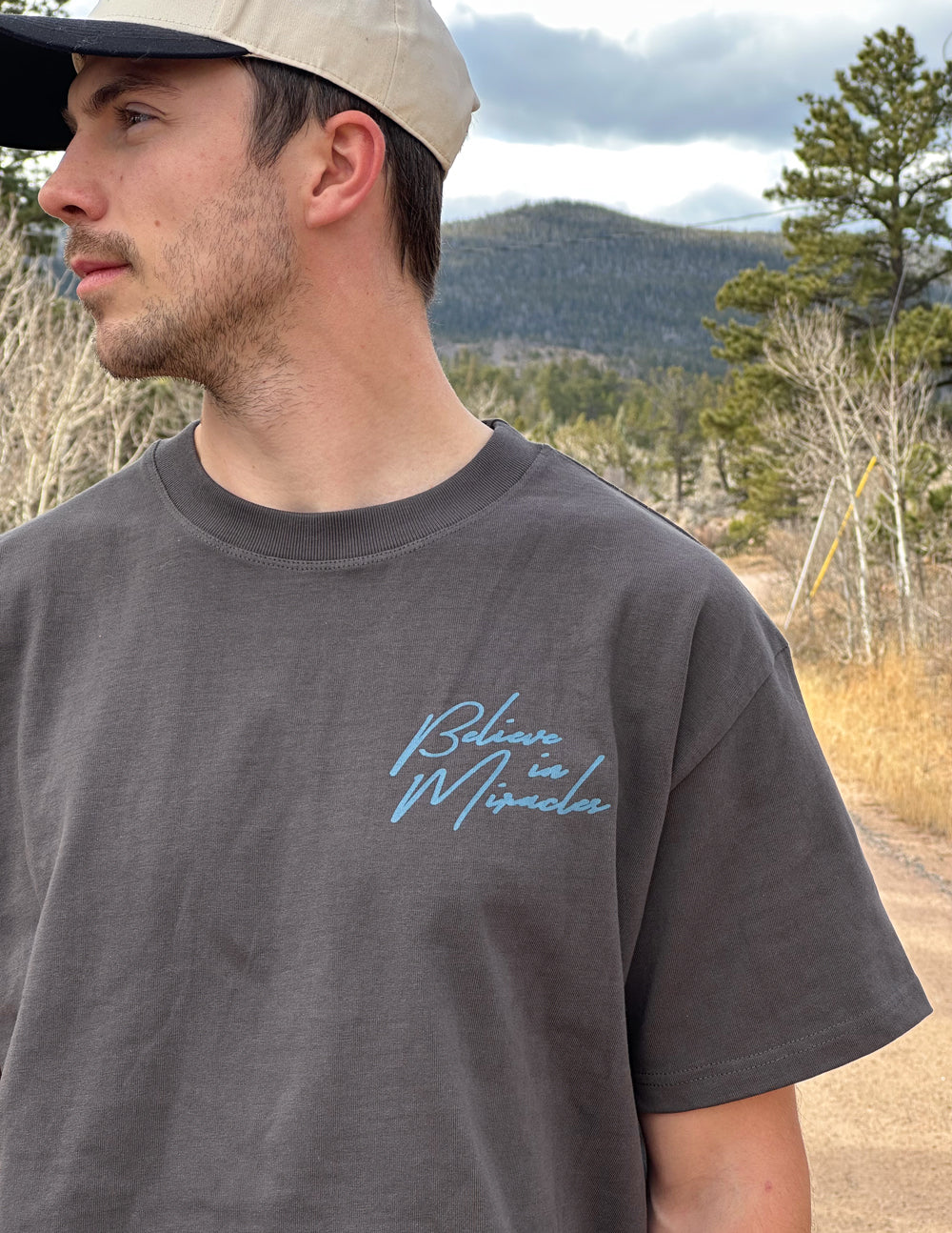 Model wearing the Lamp Black Believe in Miracles tee with a cap outdoors