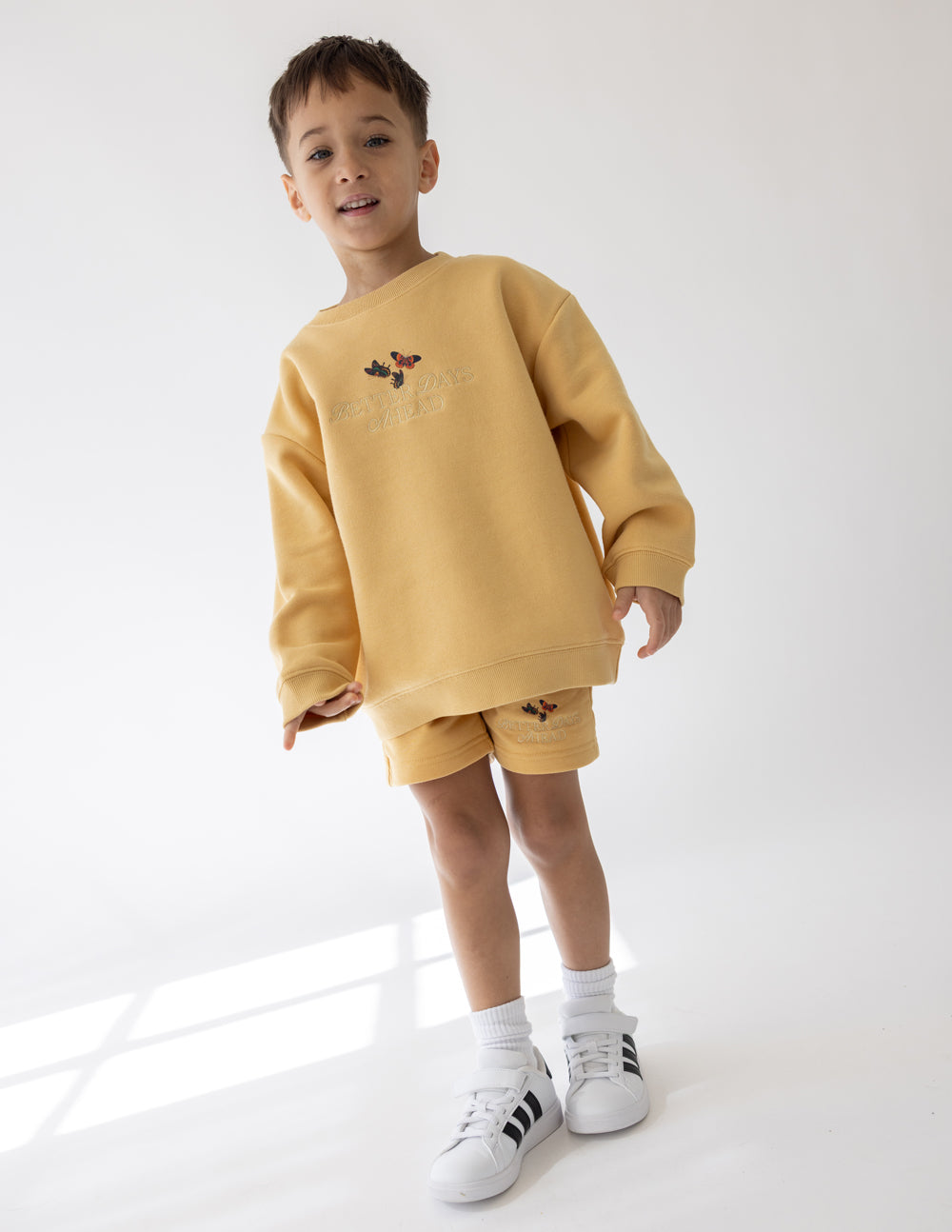 Child standing at an angle in the Better Days Butterfly crewneck sweatshirt and yellow shorts.