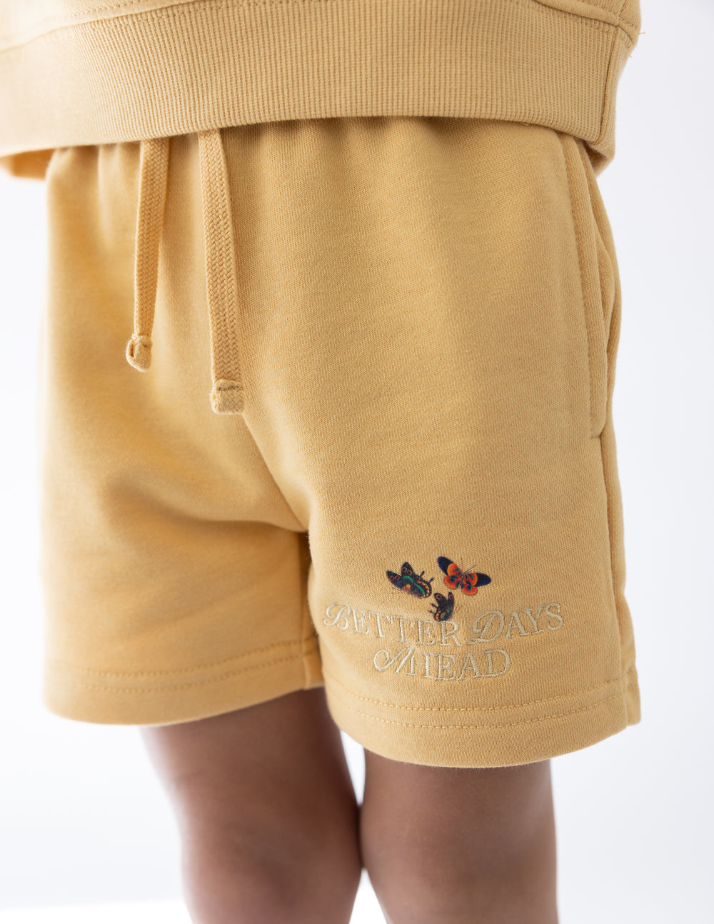 Close-up of Better Days Butterfly New Wheat kids’ shorts with a drawstring waist and embroidered butterfly design.
