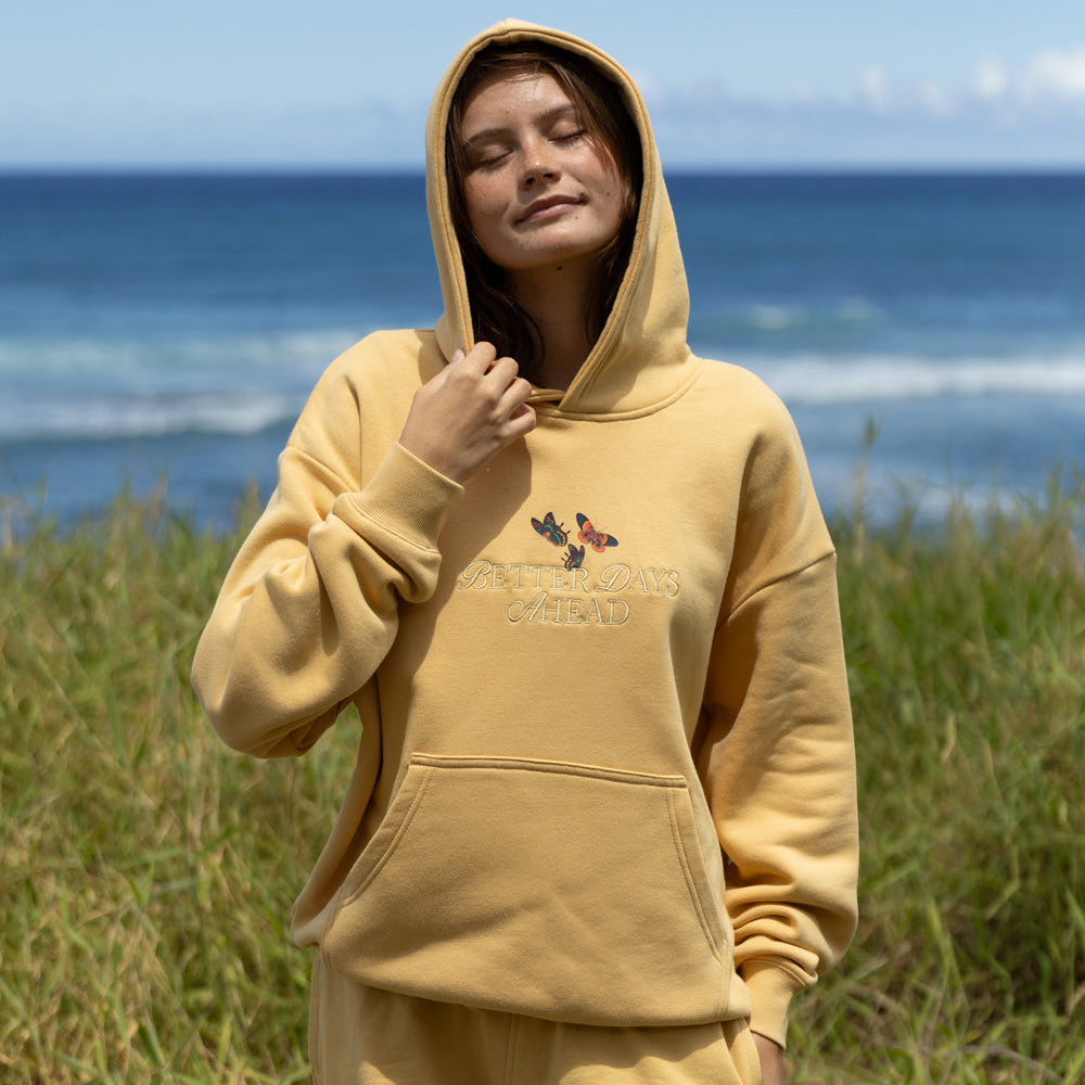 Person wearing a yellow hoodie and shorts with a graphic design, standing in a grassy area with ocean in the background