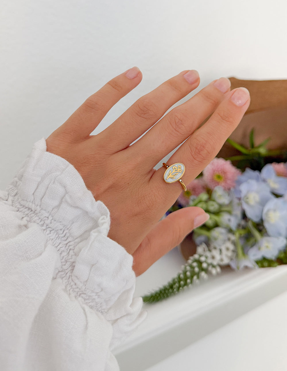 Bloom Again Enamel Ring worn on a hand beside a pastel floral arrangement