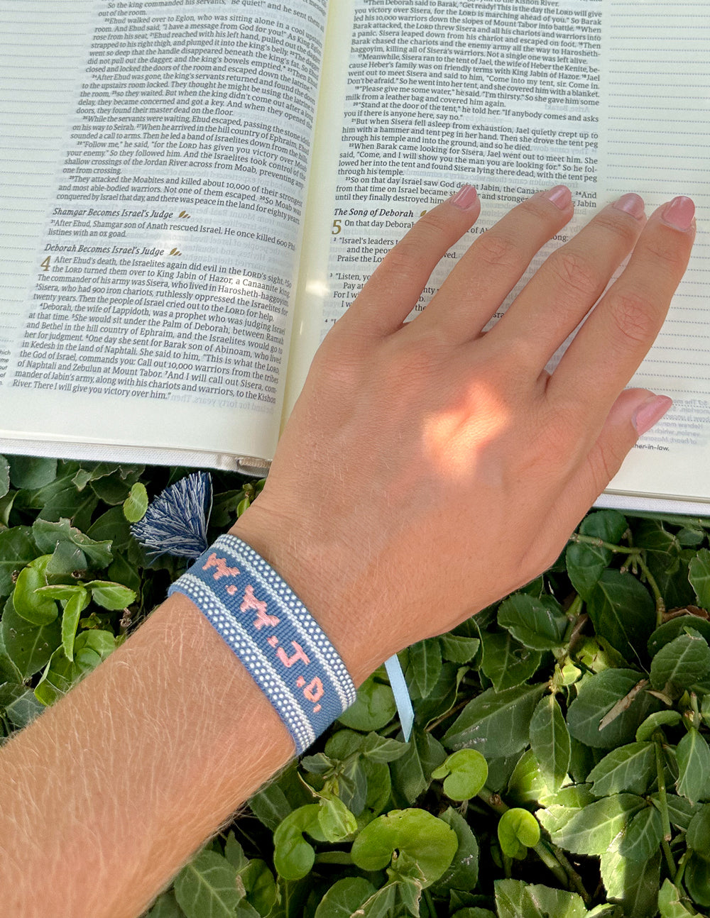 Hand wearing the blue WWJD embroidered bracelet beside an open Bible outdoors.