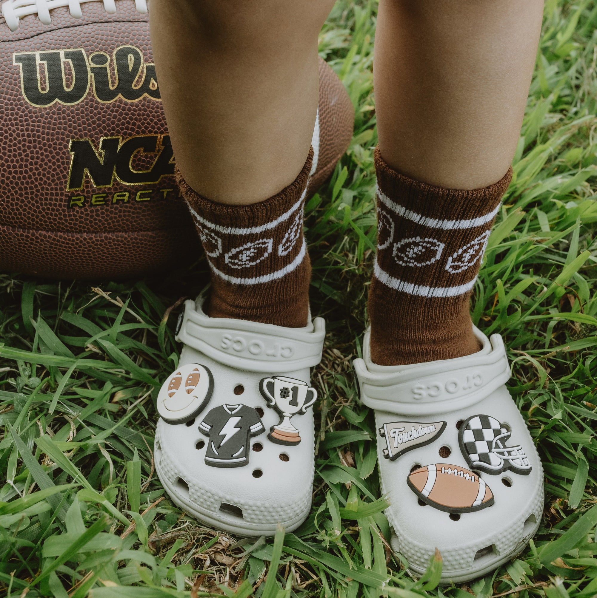 White Crocs with sports-themed designs on grass next to a Wilson NCAA football.