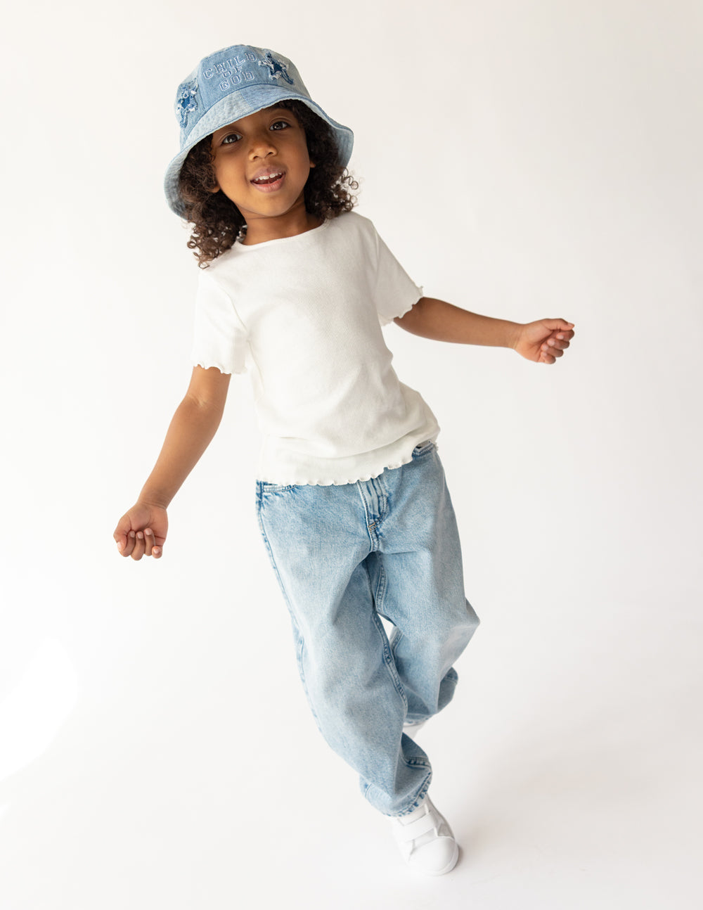 Child wearing a white t-shirt, blue jeans, and denim bucket hat on a white background