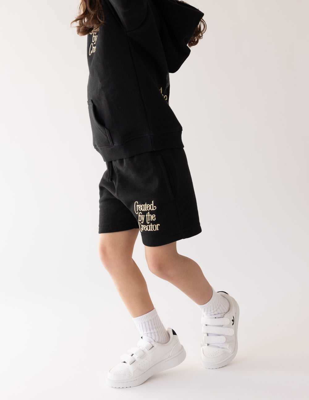 Side view of the oversized Created by the Creator Black Kids Shorts with white sneakers