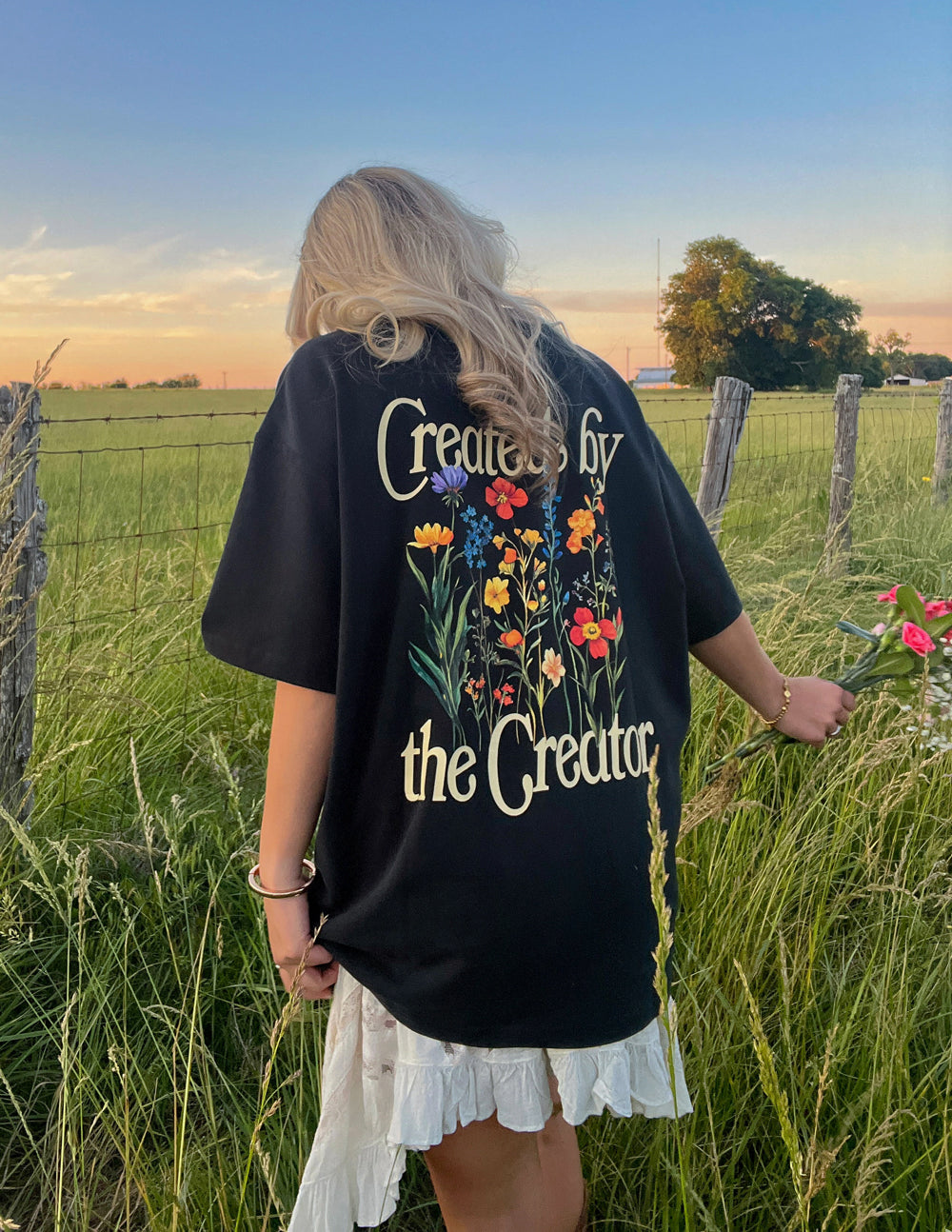 Back view of the black Created by the Creator T-shirt with colorful wildflowers and cream lettering in a grassy field.