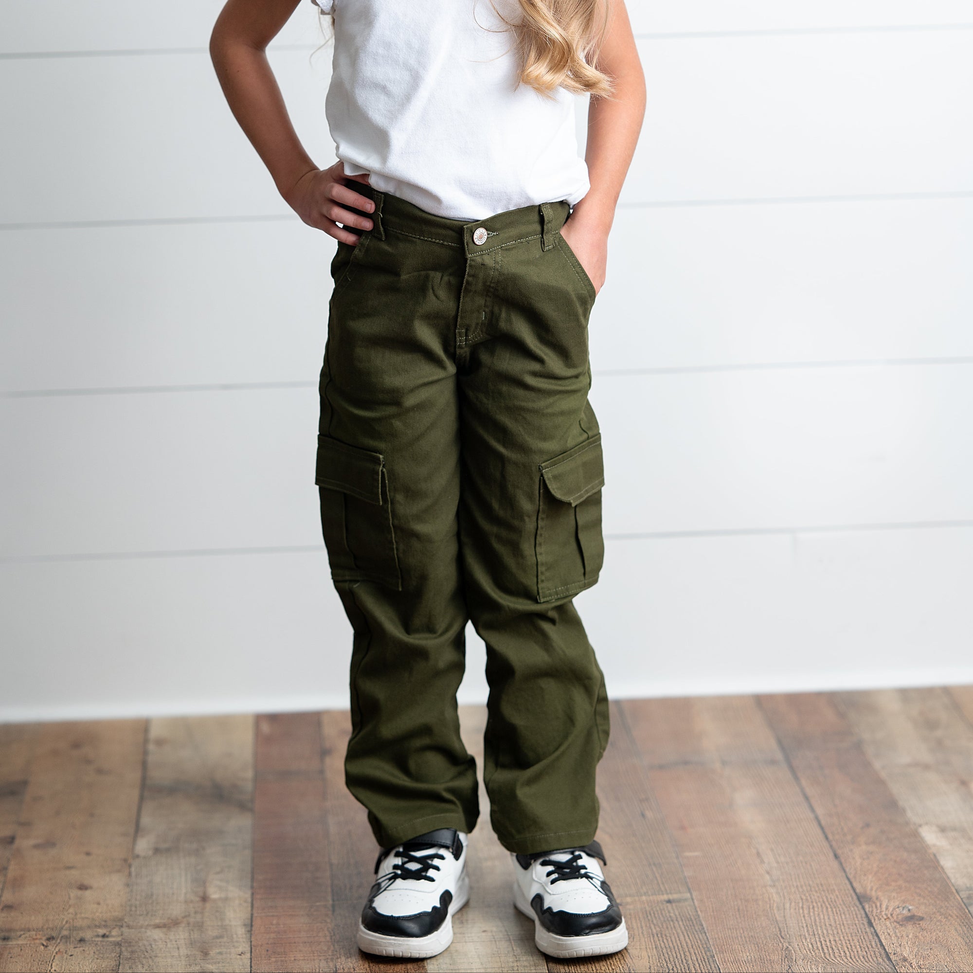 Young girl wearing a white top and green cargo pants against a white wooden panel background