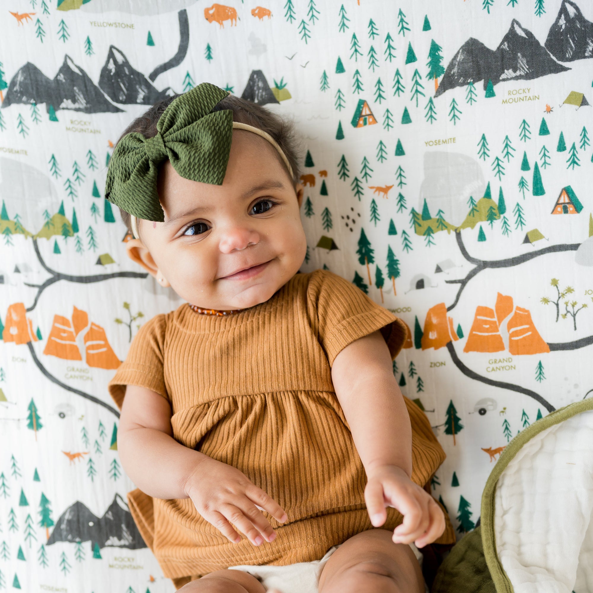 Baby in a mustard yellow outfit with a green headband sitting on a green blanket against a wall with a nature-themed wallpaper.