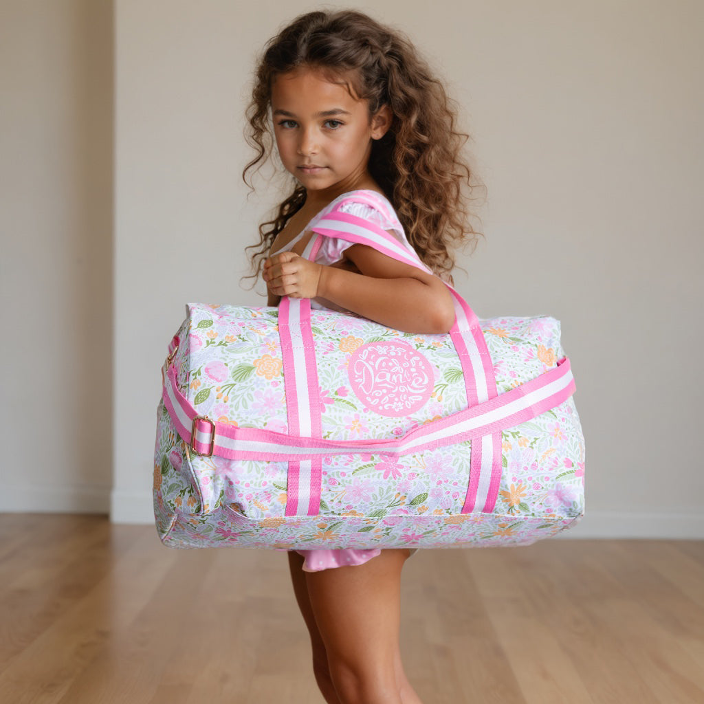 Young girl holding a floral duffel bag with pink straps on a wooden floor.