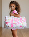 Young girl holding a floral duffel bag with pink straps on a wooden floor.