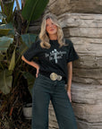 Woman styling the black cross graphic tee with dark jeans and a belt beside a stone wall.