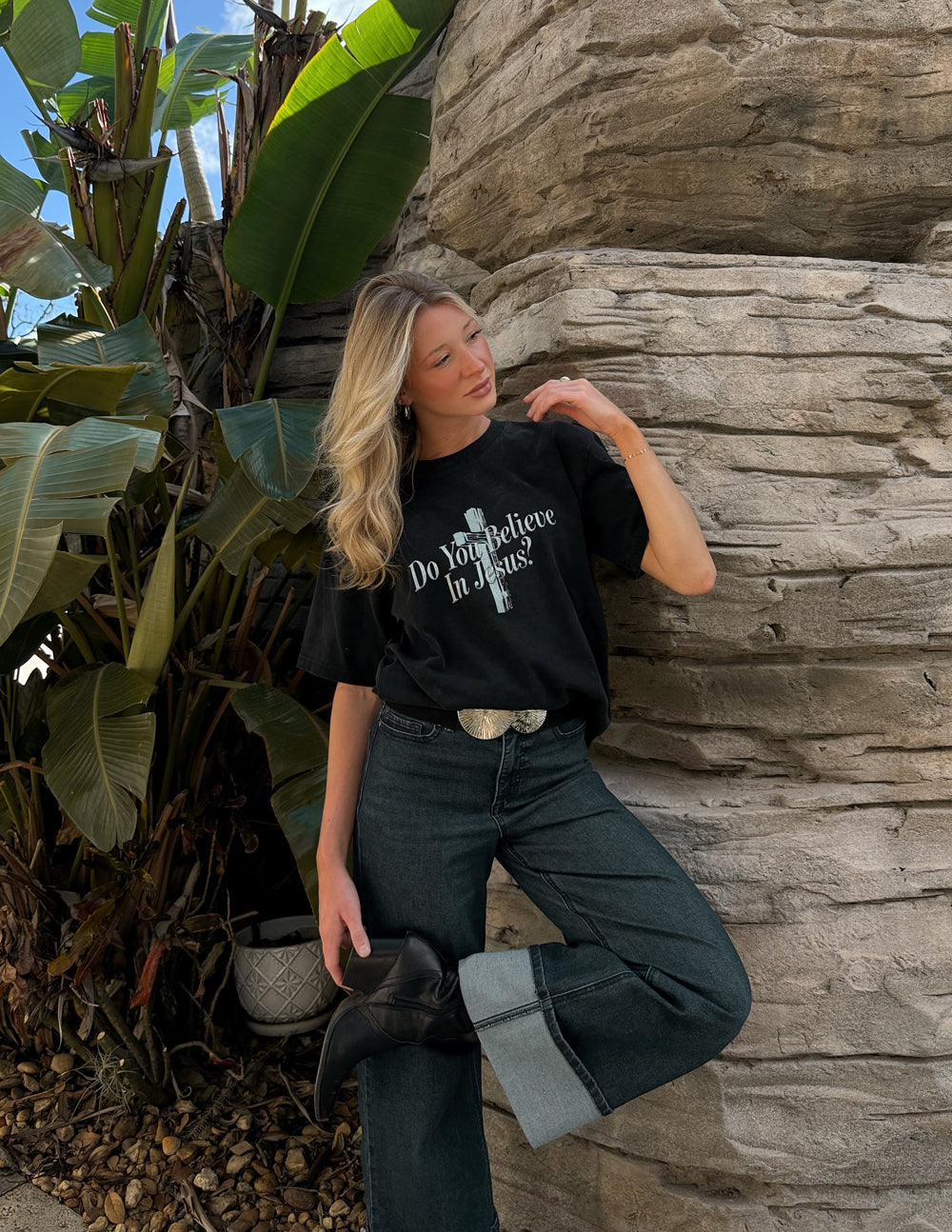 Woman posing beside a stone wall in the black Do You Believe in Jesus tee and dark jeans.