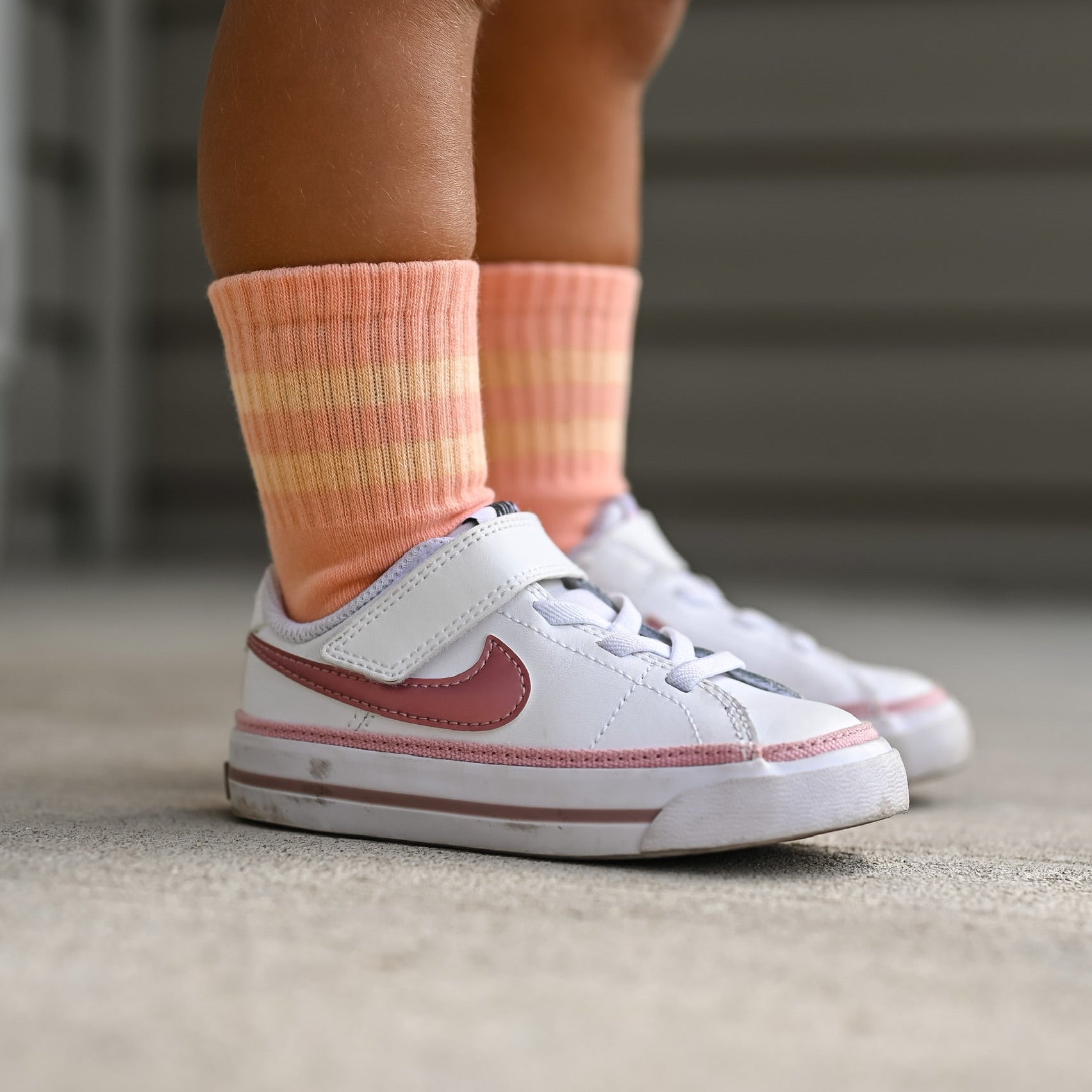 White sneakers with red accents worn by a child, paired with pink striped socks on a concrete floor.