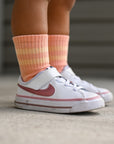 White sneakers with red accents worn by a child, paired with pink striped socks on a concrete floor.