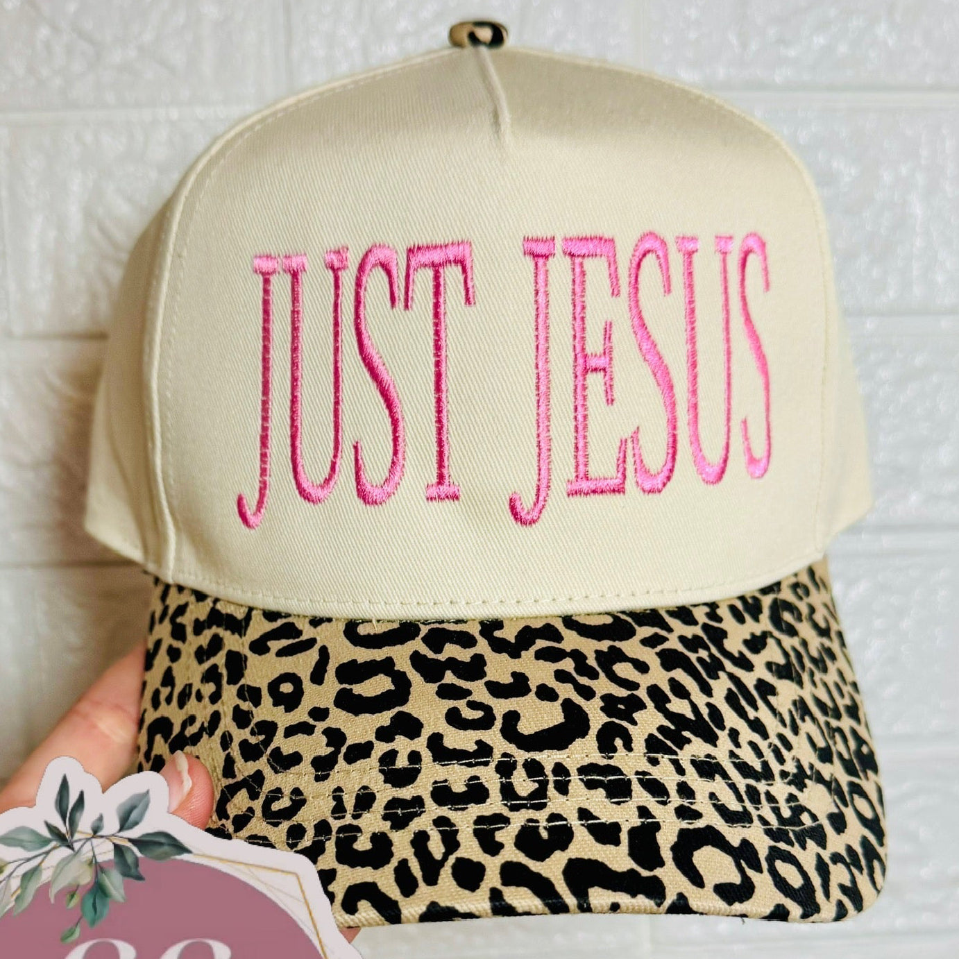 Cap with 'JUST JESUS' text and leopard print visor held by a hand, with a brand tag visible.
