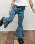 Child wearing a white top and blue jeans with a wooden floor and white wall background