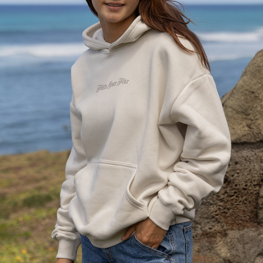 Person wearing a white hoodie with text, standing by the ocean.