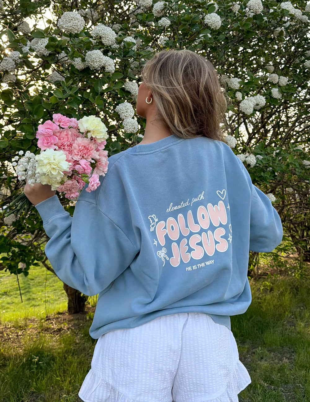 Model holding flowers while wearing the Bluebell Follow Jesus crewneck with pastel lettering on the back