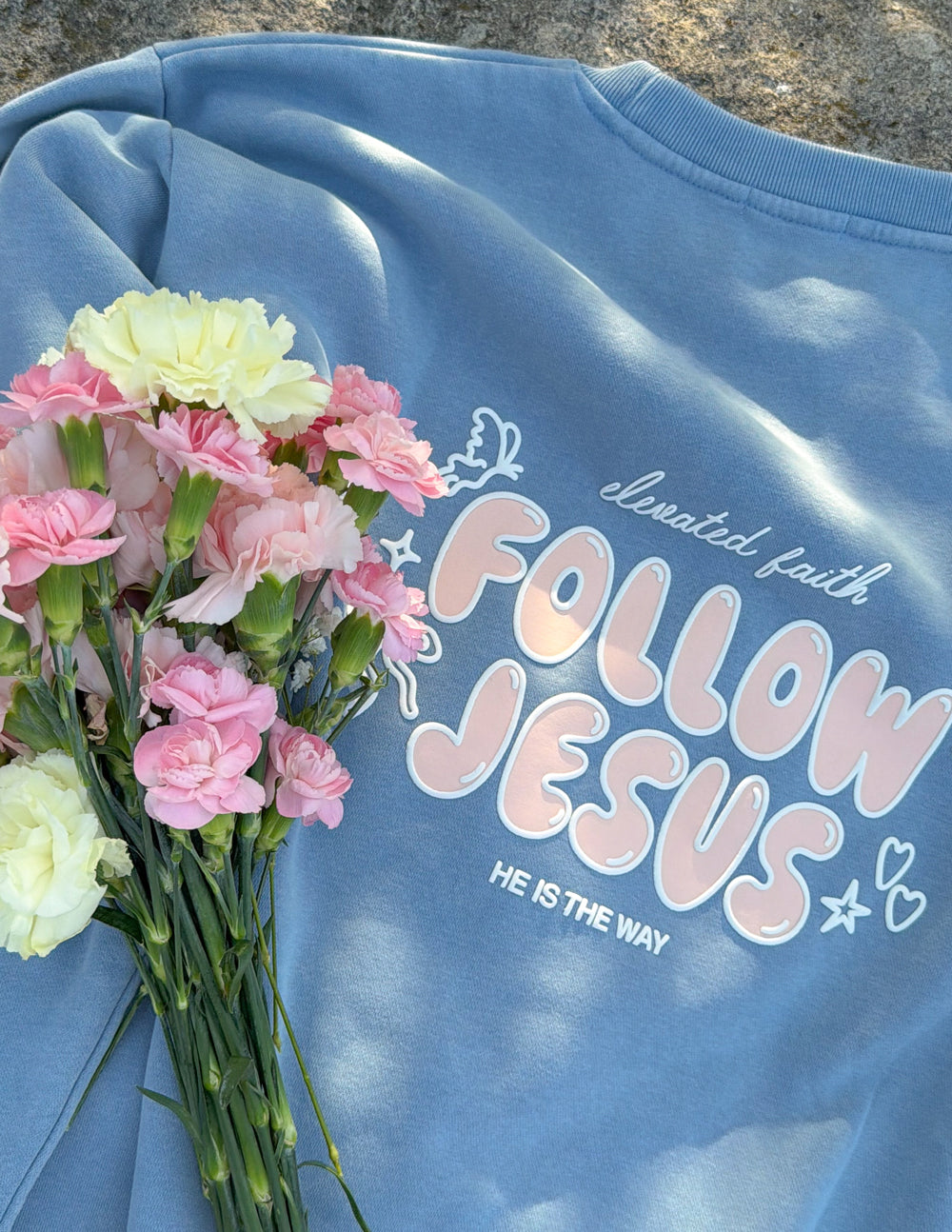Close-up of pastel Follow Jesus puff-print lettering on a Bluebell sweatshirt beside pink and white flowers