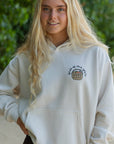 Person wearing a white hoodie with a logo, standing outdoors with greenery in the background