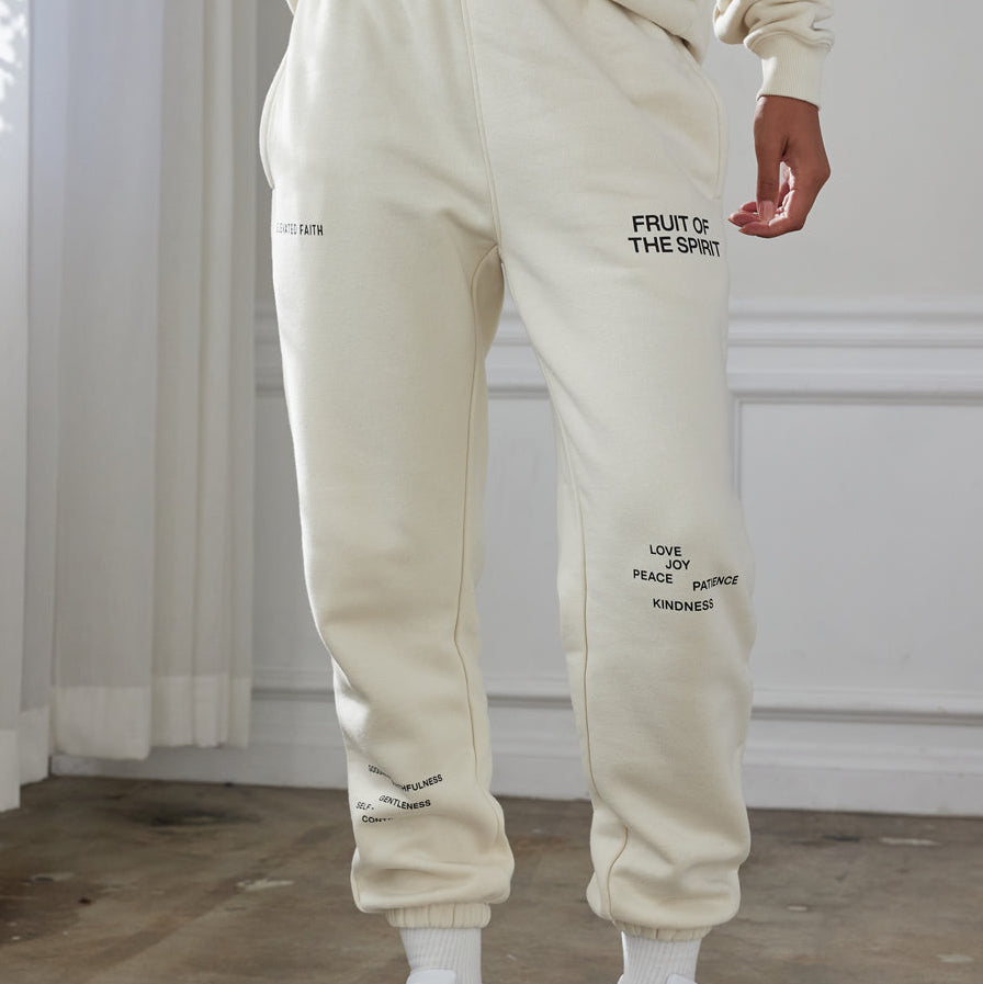 Person wearing a beige tracksuit with text on the pants, standing in a room with light-colored walls and wooden floor.