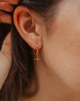 Close-up of an 18KT gold-plated cross hoop earring worn on an ear, showing the dangling cross charm