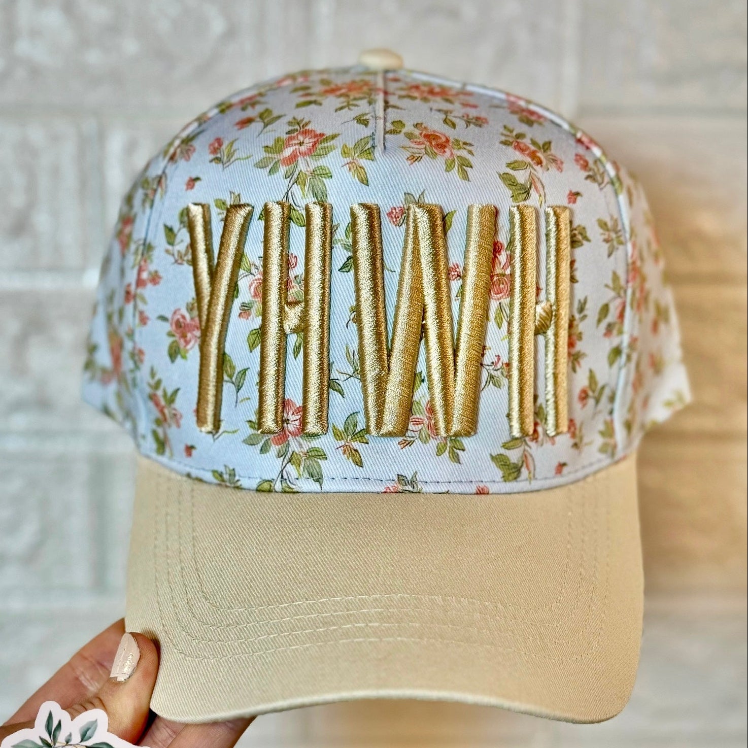 Floral-patterned cap with gold letters held by a hand, against a neutral background.