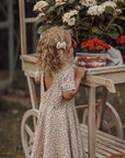 Young girl in a floral dress standing next to a wooden cart with potted plants outdoors.