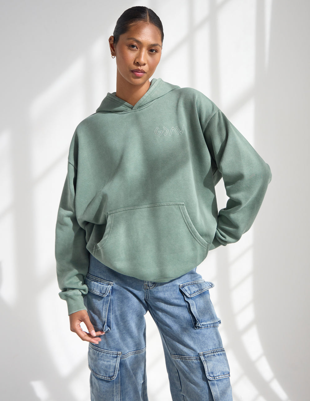 Woman wearing the moss green God Is Greater Hoodie with jeans, showing the front kangaroo pocket.
